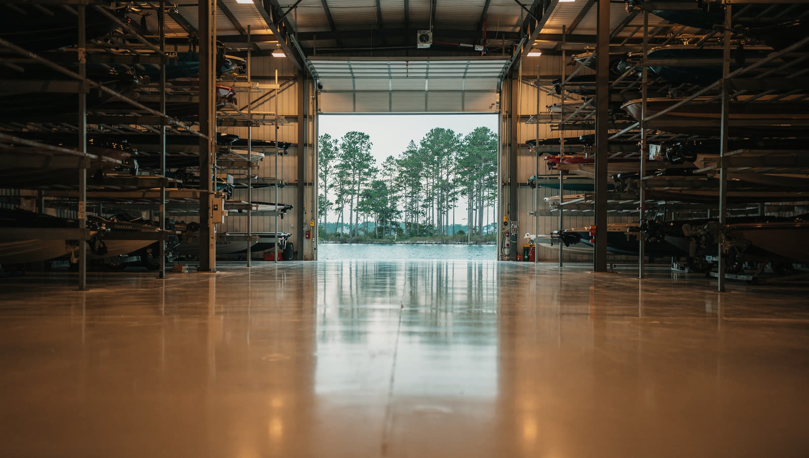 A finished commercial floor in a lakeside boat storage and service hall near Lake Conroe, tall stacked racking, loblolly pines and open water through the far door