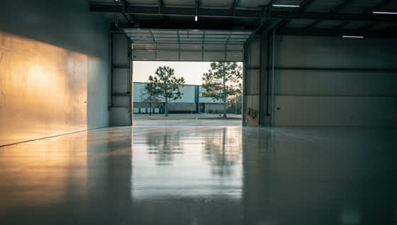 A finished commercial floor in a light industrial flex warehouse unit in a pine-lined business park, roll-up door open to a service yard